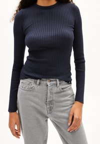 Femme portant un top rayé à manches longues bleu marine rentré dans un jean gris clair taille haute avec poches avant.