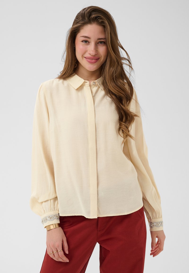 Jeune femme aux longs cheveux ondulés portant un chemisier crème avec des poignets ornés et un pantalon rouge, souriant doucement devant un fond uni.