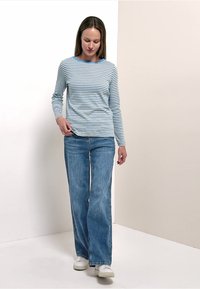 Femme portant une chemise à manches longues rayée bleu et blanc, un jean large bleu et des baskets blanches, debout devant un fond bicolore beige et blanc.