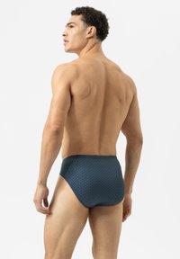 Man met krullend haar in een donkerblauw patroonondergoed, staand zonder shirt en kijkend naar links tegen een effen witte achtergrond.