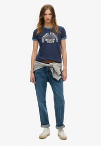 Granatowa koszulka z krótkim rękawem z białym nadrukiem "Superdry Athletics Beach Club", zestawiona z jasnoniebieskimi dżinsami z podwiniętymi nogawkami i kremowymi sneakersami. Szary sweter przewiązany w talii.