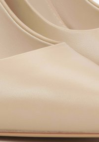 Beżowe skórzane szpilki o gładkiej teksturze, spiczastym czubku i minimalistycznym designie, z delikatnym połyskiem i bez widocznych elementów metalowych.