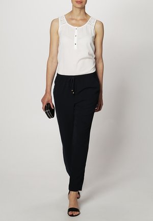 White sleeveless top with lace accents and button detail, paired with navy drawstring pants, complemented by a black clutch and sandals.