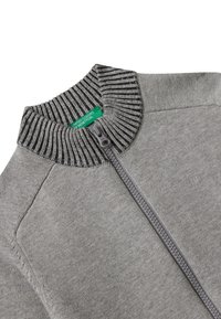 Maglione grigio con zip, caratterizzato da un colletto a righe texturizzato e una cerniera frontale. Il tag mostra "United Colors of Benetton". Design semplice e pulito.