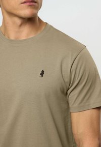 Khaki puuvillainen t-paita, jossa on pyöreä kaula-aukko ja lyhyet hihat. Vasen rinta-alue on koristeltu pienellä mustalla logolla ja t-paidassa on sileä pinta.