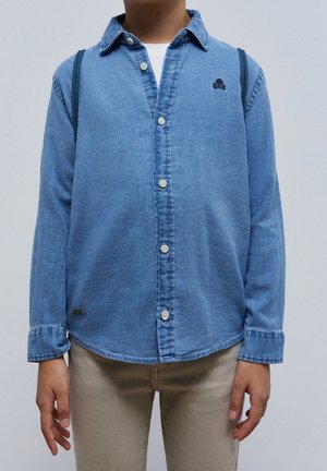 Enfant portant une chemise en denim bleu avec un petit emblème de crâne et un pantalon beige, debout devant un fond uni.