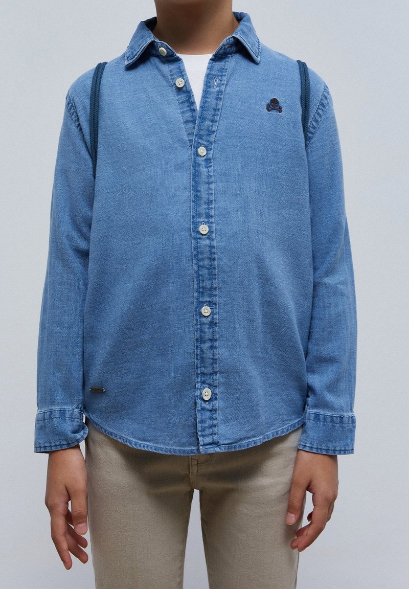 Enfant portant une chemise en denim bleu avec un petit emblème de crâne et un pantalon beige, debout devant un fond uni.