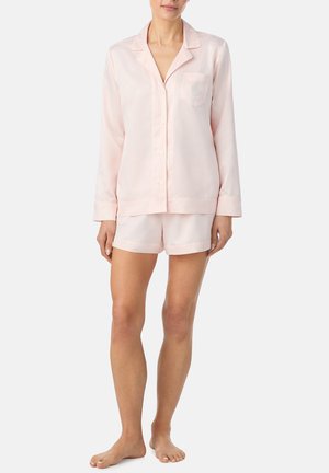 Mujer con camisa de manga larga color rosa claro abotonada y pantalones cortos a juego, de pie descalza contra un fondo liso.