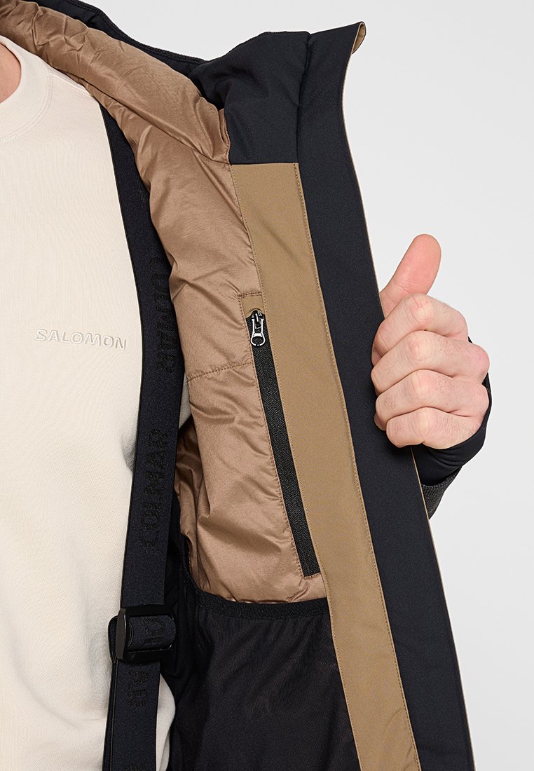 Veste isolée noire et brune avec une poche zippée et des poignets réglables, présentant une doublure intérieure douce et un tissu texturé.