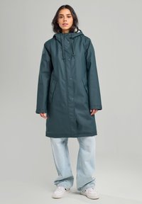 Veste de pluie teal résistante à l'eau avec capuche, boutons-pression et deux poches latérales. Associée à un jean bleu clair à jambes larges et des baskets blanches.