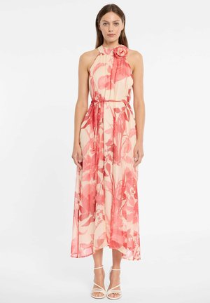 Frau steht und trägt ein ärmelloses Maxikleid mit Neckholder, rosa und cremefarbenem Blumenmuster, mit einem Blumenakzent auf der Schulter und weißen Riemchenabsätzen.
