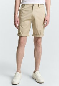 Beige Shorts mit umgeschlagenen Säumen aus Baumwollmischgewebe, mit einem Knopfverschluss und flachem Schnitt, kombiniert mit weißen Sneakers.