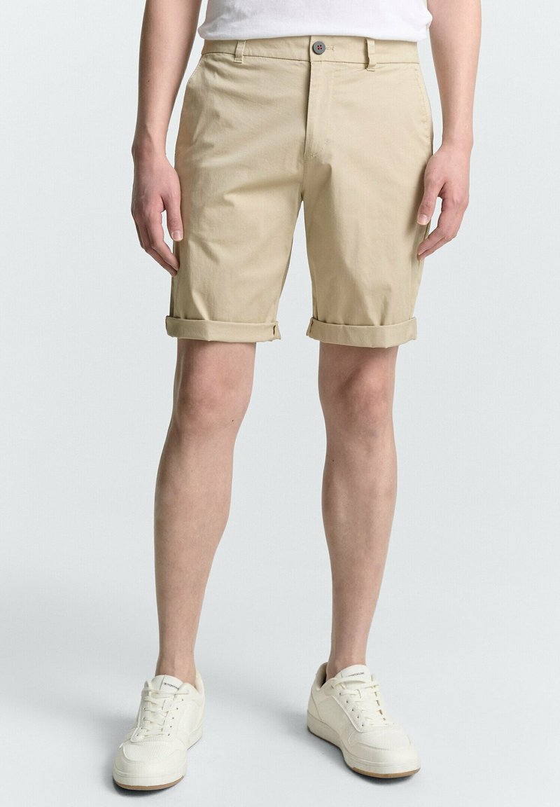 Beige Shorts mit umgeschlagenen Säumen aus Baumwollmischgewebe, mit einem Knopfverschluss und flachem Schnitt, kombiniert mit weißen Sneakers.