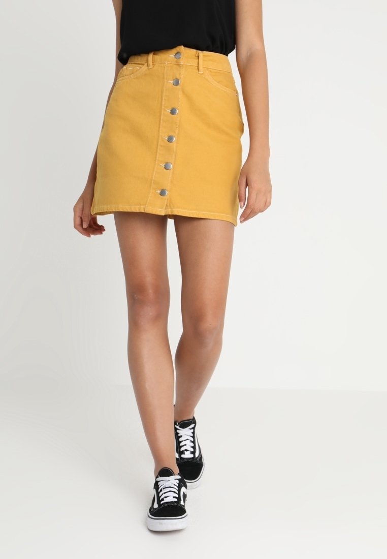 Personne portant une mini-jupe en jean jaune moutarde avec boutons sur le devant, un haut sans manches noir et des baskets montantes noires et blanches.