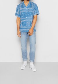 Camicia blu a maniche corte con bottoni e disegni a fantasia, abbinata a jeans azzurri e sneaker bianche alte.