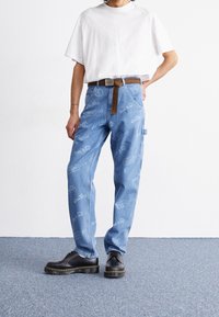 Hellblaue Denim-Hosen mit lockerer Passform, ausgestattet mit wiederholten weißen Logo-Mustern. Kombiniert mit einem weißen Hemd und schwarzen Schuhen. Brauner Gürtel als Akzent.