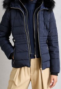 Veste matelassée bleu marine avec capuche en fausse fourrure, fermeture éclair à l'avant et poches latérales. Présente des coutures horizontales et des détails en métal argenté.
