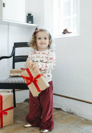 Ein Kind hält ein eingewickeltes Geschenk mit einem roten Band. Sie trägt ein langärmliges Shirt mit Schleifenmustern und weinrote Hosen mit Rüschchen.