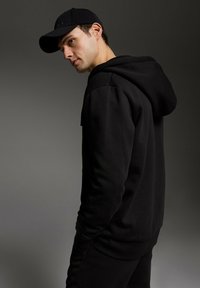 Czarna bluza z kapturem z kieszenią na przodzie i sznurkiem do regulacji, wykonana z teksturowanej tkaniny. Noszona z pasującą czarną czapką, podkreślającą sportowy styl.