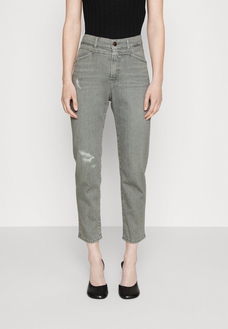 Femme portant un jean gris taille haute légèrement déchiré, un top noir sans manches et des chaussures à talons noires, debout devant un fond uni.