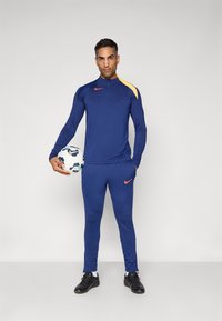 Nike sporta apģērbs tumši zils ar dzeltenu plecu detaļu, ietverot rāvējslēdzēja apkakli un pieguļošas bikses, kopā ar futbola bumbu.