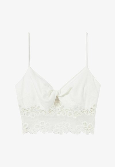 Crop top blanc avec des bretelles fines ajustables, présentant un détail de nœud à l'avant et une bordure en dentelle florale à l'ourlet. Tissu doux et lisse.