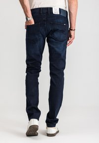 Mężczyzna w ciemnoniebieskich dżinsach i białych tenisówkach, z jedną ręką w tylnej kieszeni, widoczny od tyłu na jednolitym tle.