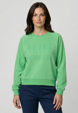 Frau mit dunklen Haaren, die ein grünes Sweatshirt mit dem Schriftzug "ALIFE WE ARE ALIFE" und eine marineblaue Hose trägt, stehend vor einem schlichten Hintergrund.