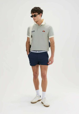 Man draagt een grijs poloshirt, donkerblauwe korte broek, witte sneakers, zonnebril en een schoudertas, staand tegen een effen witte achtergrond.