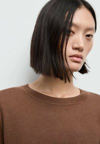 Brun strikket sweater med rund halsudskæring og fint mønster, præsenteret mod en neutral baggrund. Korte mørke hår, minimal makeup.