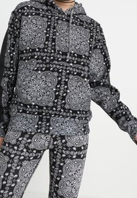 Sudadera con estampado de bandana en negro y gris, con un bolsillo delantero y capucha, combinada con mallas a juego que presentan un patrón intrincado similar.