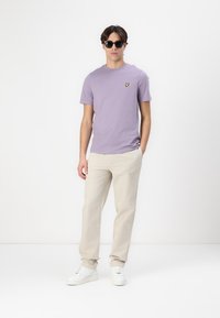 T-shirt en coton lilas avec un petit logo jaune sur la poitrine, associé à un pantalon couleur crème et des baskets blanches. Le mannequin porte des lunettes de soleil noires.