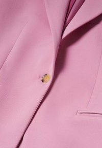 Gros plan sur un blazer rose clair avec un seul bouton beige et une poche passepoilée étroite sur un tissu lisse.
