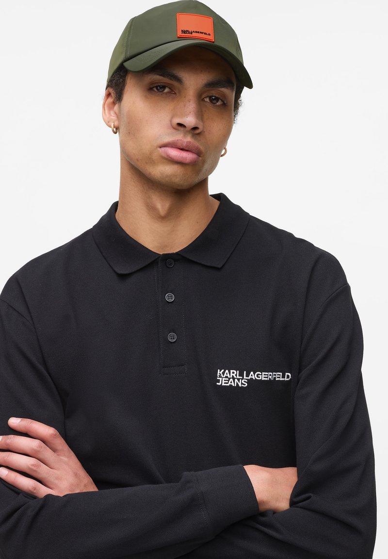 Fekete póló ing három gombbal és hímzett "KARL LAGERFELD JEANS" logóval. Durva szövettextúra. Olívazöld sapka négyzet alakú narancssárga címkével.
