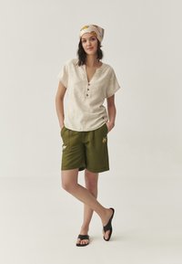 Lichtbeige korte mouw top met bloem broderie en knopen; olijfgroene shorts met dierenpatches; zwarte slippers.