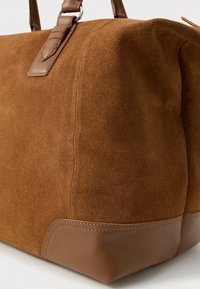 Bruine nubuck en leren reistas met ronde hoeken en gestikte leren handvatten, weergegeven in close-up op een witte ondergrond.