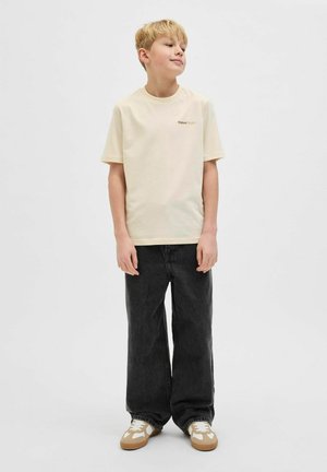 Hellbeige Baumwoll-T-Shirt mit kurzen Ärmeln und Logodetails, kombiniert mit locker sitzenden schwarzen Jeans und weißen Sneakers mit beigen Akzenten.