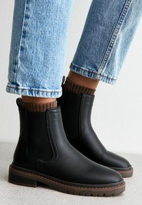 Botines Chelsea de cuero negro con paneles laterales elásticos, punta redondeada y detalle de calcetín marrón texturizado. Suela marrón plana y resistente.