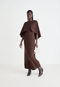 Vestido largo marrón, texturizado, con una parte superior de capa fluida, combinado con botas de tacón grises y un pequeño bolso blanco. Diseño simple y aerodinámico.
