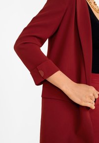 Blazer bordeaux avec une texture lisse, doté d'un poignet replié et d'un accent circulaire. Stylé avec un haut noir et une jupe assortie.