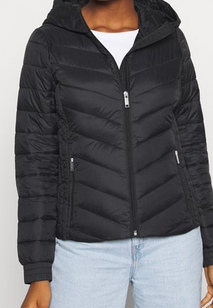 Veste noire matelassée à fermeture éclair avec capuche, portée par-dessus un haut blanc et un jean bleu clair, vue des épaules aux hanches.