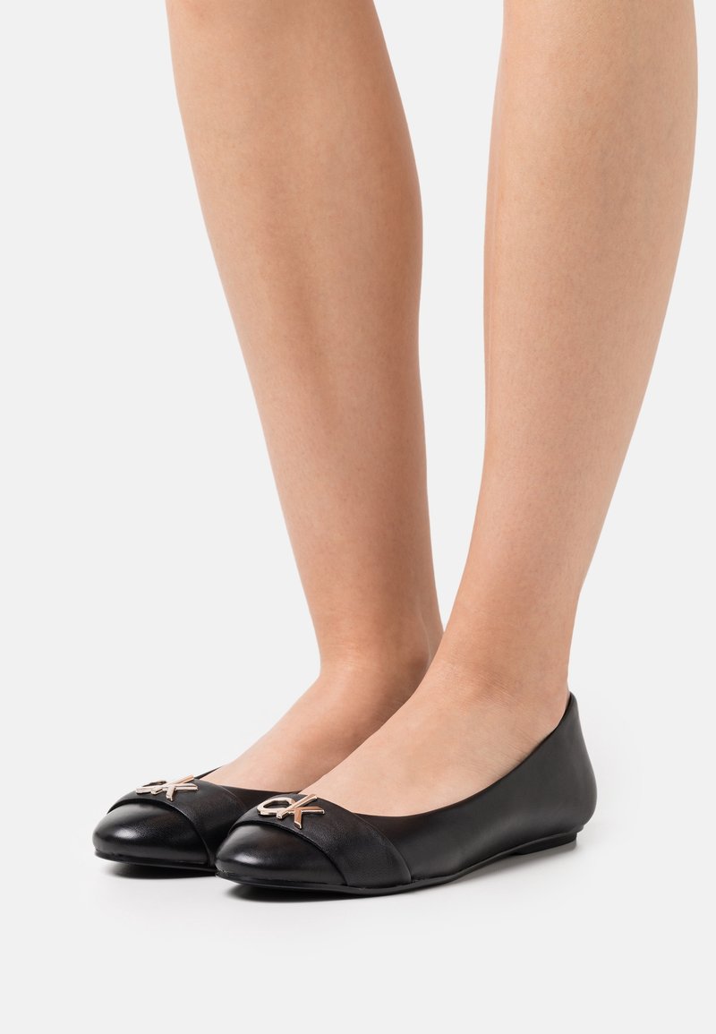 Jambes portant des ballerines en cuir noir à bouts pointus avec des ornements métalliques du logo Calvin Klein sur fond blanc.