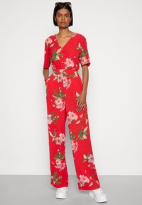 Macacão floral vermelho com mangas curtas, frente com botões, cintura elástica, pernas largas, e flores cor-de-rosa com folhas verdes, emparelhado com ténis brancos.