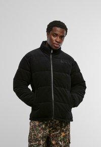 Mustast korduroost pufferjakk kõrge krae, luku ja tikandmustriga. Rinnal silmatorkav õmblusdetail. Vabaaja stiil.