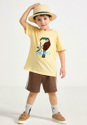 Nuori poika, joka käyttää olkihattua, keltainen t-paita, jossa on tukaani-kuvio, ruskeat shortsit, tukaani-kuvioidut sukat ja ruskeat tarrakengät.