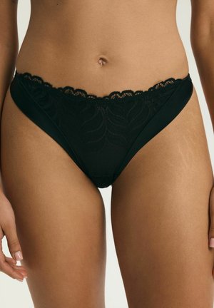 Kobieta w czarnym bikini z koronkowymi wykończeniami, pokazująca środkową część ciała i górną część ud na jednolitym tle.