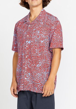 Mann trägt kurzärmliges Hemd mit roten abstrakten Mustern auf hellblauem Hintergrund und dunkle Hose.