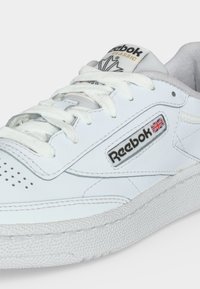 Zapatilla blanca Reebok Classic con cordones blancos, logo visible de Reebok y pequeña bandera del Reino Unido en el lateral.
