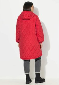Red quilted long coat with a hood, featuring a diamond pattern. Worn over gray trousers and black ankle boots. Soft texture and casual design.