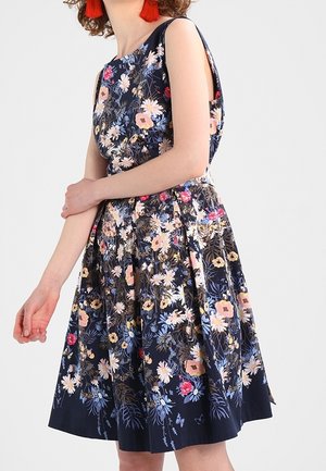 Kobieta w granatowej sukience bez rękawów, o długości do kolan, z wielokolorowym kwiatowym wzorem, nosząca czerwone kolczyki z chwostami, stoi z ramionami skrzyżowanymi.